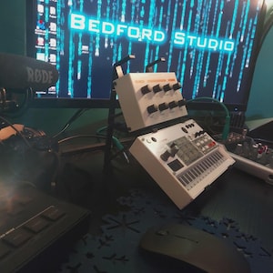 May include: A home studio setup with a RODE microphone, a computer monitor displaying "BEDFORD STUDIO", and various audio equipment. The setup includes a synthesizer and a mixing board, all on a desk.