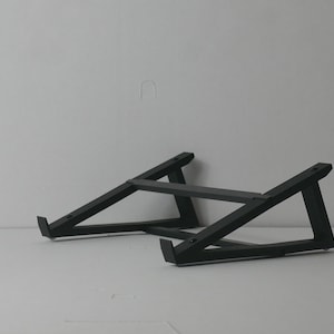 Puede incluir: Un soporte negro para portátil con un diseño minimalista. El soporte está hecho de soportes angulados y resistentes y una barra horizontal, creando una base estable. Diseñado para elevar un portátil.