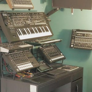 Puede incluir: Una configuración de estudio de música con múltiples sintetizadores, teclados y mezcladores de audio negros. El equipo está montado en soportes y un escritorio contra una pared verde azulado. También hay un teclado de computadora y un monitor.