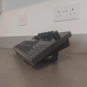 May include: A gray audio mixing console with numerous knobs, sliders, and buttons. The device is angled upwards, supported by a built-in stand. The console is placed on a light gray surface, with white electrical outlets in the background.