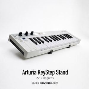 May include: A white Arturia KeyStep stand with black keys and control knobs. The stand is angled at 22.5 degrees and has the text "Arturia KeyStep Stand" and "studio-solutions.com" printed on it.
