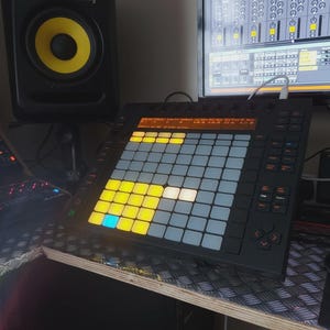 Pode incluir: Dispositivo preto de produção de música eletrónica com uma grelha de pads iluminados em amarelo, azul e branco. Uma grande coluna com um centro amarelo está no fundo. Um monitor de computador exibe uma interface de produção musical.