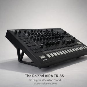 Puede incluir: Batería electrónica Roland AIRA TR-8S negra sobre un soporte de escritorio de 30 grados. El instrumento musical electrónico presenta numerosas perillas, botones y un teclado. El soporte es negro y angulado.