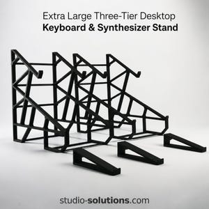 May include: An extra large three-tier desktop keyboard and synthesizer stand in black. The structure features a geometric, open-frame design with multiple support arms for holding musical equipment. Text reads: Extra Large Three-Tier Desktop Keyboard & Synthesizer Stand and studio-solutions.com.