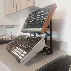 May include: A black metal stand supporting two electronic music synthesisers. The top synth has a wooden side panel. The bottom synth is black and silver. The word "ARTURIA" is visible on the top synth.