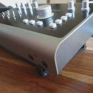 May include: A silver and black audio interface with multiple knobs, buttons, and input jacks. The device has a sleek, modern design with a matte finish. The top panel features various controls, including faders and rotary knobs. The unit is designed for professional audio recording and mixing.