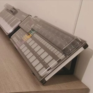 Puede incluir: Un dispositivo gris de producción de música electrónica con un teclado y múltiples pads, cubierto por un escudo de plástico transparente. El dispositivo está inclinado sobre una superficie de madera, con una pared blanca en el fondo.