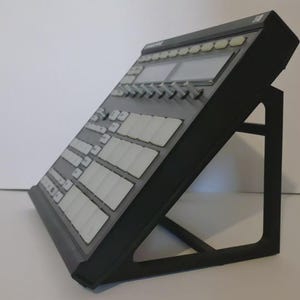 May include: A black electronic music production device, featuring buttons, pads, and screens, mounted on a black stand. The device is angled for ease of use. It is designed for music production and performance, with a sleek, modern design.
