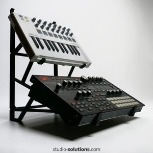 May include: A black metal stand holding two electronic music production devices. The top device is a white Arturia Minilab with black keys and knobs. The bottom device is black with red accents and numerous knobs and buttons. The stand is angled.