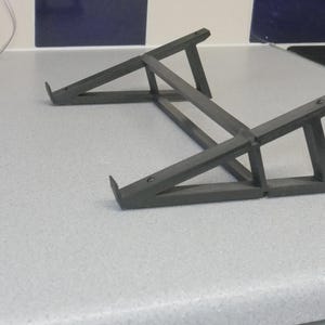 Op de afbeelding: Een donkergrijze laptopstandaard met een geometrisch ontwerp. De standaard is gemaakt van stevig materiaal en heeft schuine steunen voor stabiliteit. Het is ontworpen om een laptop te verhogen.