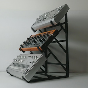 Może przedstawiać: Czarny metalowy stojak z trzema srebrnymi i pomarańczowymi syntezatorami muzyki elektronicznej. Stojak ma geometryczny kształt, a syntezatory są ustawione pod kątem. Idealny do studia muzycznego.