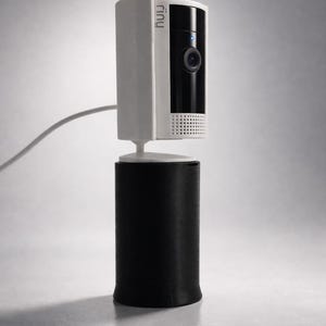 Puede incluir: Una cámara Ring Doorbell 2 blanca y negra montada sobre un elevador cilíndrico negro. La cámara tiene una lente y una pequeña luz azul. Se ve el texto "Ring Doorbell 2" y "Riser".