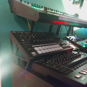May include: A collection of black electronic music production devices, including a keyboard and several control surfaces. The keyboard is mounted above the other devices. The devices have various buttons, knobs, and sliders. The scene is lit with green and red lights.