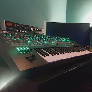 May include: A black synthesizer with a keyboard and various knobs and buttons. The synthesizer has green illuminated controls and a white and black keyboard. Another piece of equipment is stacked on top of the synthesizer.