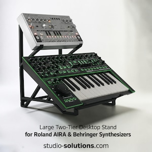 以下が含まれることがあります： 上段にシルバーのBehringer TD-3シンセサイザー、下段に黒と緑のRoland System-1シンセサイザーを載せた黒い2段式デスクトップスタンド。テキストには「Large Two-Tier Desktop Stand for Roland AIRA & Behringer Synthesizers」および「studio-solutions.com」と記載されています。