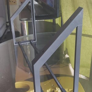 Op de afbeelding: Een zwarte, geometrische framestructuur met een helder glazen paneel, rustend op een goudkleurige gitaarvormige basis. Het frame heeft een modern ontwerp, met schuine steunen. De achtergrond omvat een tv en een potplant.