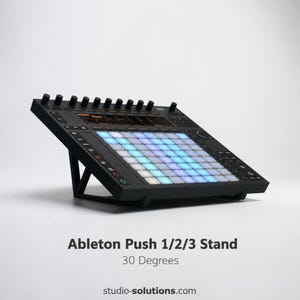 May include: A black Ableton Push 1/2/3 stand angled at 30 degrees. The stand supports a music production controller with a grid of illuminated pads in blue, teal, and white. The stand has a minimalist design and is made of black metal.