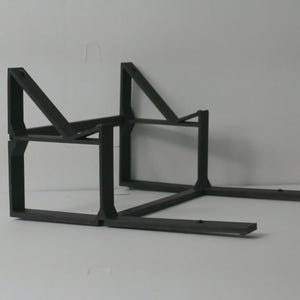 May include: A dark gray, geometric, 3D-printed stand. The stand has a rectangular base with two parallel arms extending forward. Two vertical supports rise from the base, connected by angled supports, creating a sturdy structure.