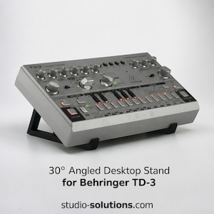 Puede incluir: Un sintetizador analógico Behringer TD-3 de color plateado sobre un soporte de escritorio negro inclinado a 30 grados. El sintetizador cuenta con numerosos mandos, botones y una interfaz de teclado con luces naranjas en un panel gris.