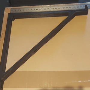 Puede incluir: Un soporte de estante en ángulo recto, de color marrón oscuro. El soporte está hecho de un material sólido y presenta una viga de soporte diagonal. Una regla metálica se coloca encima del soporte, mostrando medidas en centímetros.