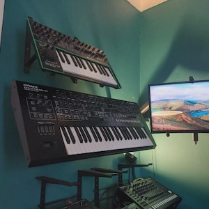 May include: Two synthesizers mounted on a teal wall, one black and one green. The black synthesizer is labelled "Roland SYSTEM-100". A television displays a landscape scene. A mixing console sits below the synthesizers.