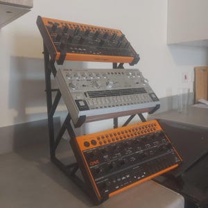 Op de afbeelding: Een zwart metalen statief met drie synthesizers. De bovenste en onderste synthesizers zijn oranje en zwart, de middelste zilverkleurig. Het statief is schuin geplaatst, met de merken Behringer en Dave Smith Instruments.