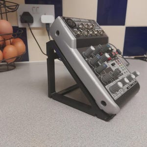 May include: A silver and black audio mixer with numerous knobs and input jacks, positioned on a black stand. The mixer is on a speckled countertop, with a wire basket of brown eggs in the background.