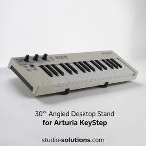 May include: A white 30-degree angled desktop stand supporting an Arturia KeyStep MIDI keyboard controller. The keyboard features white and black keys, three black rotary knobs, and various control buttons. Text on the image reads 30-degree Angled Desktop Stand for Arturia KeyStep and studio-solutions.com.