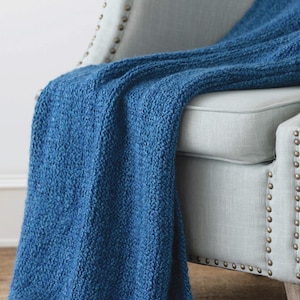 Puede incluir: Una manta de punto azul texturizada sobre un sillón gris claro. El color intenso de la manta contrasta con el tono neutro de la silla. La silla tiene un marco de madera clara y está adornada con tachuelas decorativas.