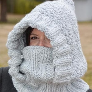 May include: A light gray knitted hood with a ribbed cowl neck. The hood is made of a thick, textured yarn and has a wide, face-framing design. The cowl neck is pulled up to cover the lower face. The hood is worn over a dark gray coat.