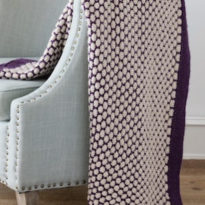 May include: A cream and purple knitted throw blanket draped over a light blue armchair. The blanket features a textured pattern of cream-colored circles against a purple background. The blanket's edge is a solid purple color.