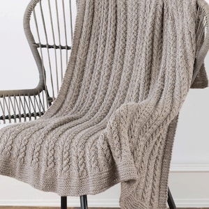 Puede incluir: Una manta de punto marrón claro con un diseño de punto de cable está sobre una silla de mimbre gris. La manta tiene un aspecto texturizado con patrones verticales y diagonales. La silla tiene respaldo curvo y patas de metal negro.