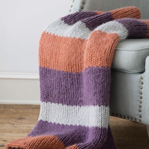 Puede incluir: Una manta tejida a mano con amplias rayas horizontales en tonos naranja, morado y blanco, colocada sobre un sillón azul claro. La gruesa textura de punto sugiere calidez y comodidad. Un elemento de decoración para el hogar.