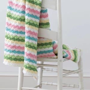 Puede incluir: Una manta de punto a rayas multicolores, compuesta por bandas horizontales blancas, rosas, amarillas, verdes y azules, sobre una silla de madera blanca. El patrón texturizado añade interés visual al textil suave y acogedor.