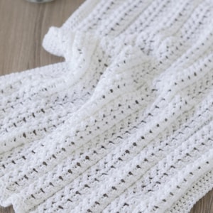 Puede incluir: Una manta blanca de ganchillo hecha a mano con un diseño de tejido abierto y texturizado. La manta está doblada, mostrando el intrincado patrón de puntadas y la textura suave. El color es un blanco brillante y limpio, ideal para un bebé o decoración del hogar.