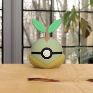 Puede incluir: Una bola decorativa verde, amarilla y marrón con dos hojas verdes en la parte superior. La bola tiene una franja negra y un círculo blanco en el centro. Está sobre una superficie de madera.
