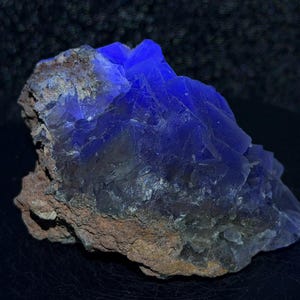 Puede incluir: Un espécimen mineral en bruto que presenta estructuras cristalinas de color azul translúcido vibrante que sobresalen de una matriz rocosa de color marrón tierra rugoso. El grupo muestra facetas geométricas afiladas que captan la luz, contrastando con la base texturizada.