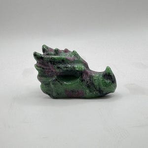 May include: A carved dragon head figurine. The sculpture is made of a green and maroon stone. The dragon's head is detailed with textured scales and a pointed snout. The figurine is set against a white background.
