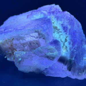 Puede incluir: Un espécimen de mineral de fluorita en bruto que exhibe una intensa fluorescencia azul bajo luz ultravioleta. La estructura cristalina presenta formaciones cúbicas con tonos púrpuras y azules translúcidos, acentuados por inclusiones minerales blancas internas.