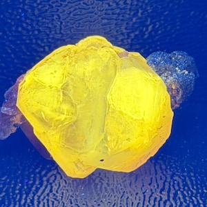 Puede incluir: Un espécimen mineral amarillo brillante y translúcido con una estructura cristalina multifacética. El grupo amarillo central está unido a una formación rocosa más pequeña, oscura y texturizada, creando un marcado contraste de color.