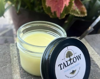 May include: A small glass jar filled with yellow baby butter, with the lid off to the side. The lid is black with a white label that reads "TALLOW Healing Baby Butter" with a tree and compass design. The jar sits on a brick surface.