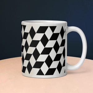 May include: White ceramic mug with a black and white geometric diamond pattern. The mug has a curved handle and is sitting on a light pink surface. The background is a dark blue.
