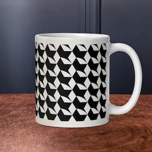 May include: White ceramic mug with a repeating black and white geometric pattern. The pattern consists of interlocking shapes, creating a visually striking design. The mug has a curved handle and is set against a dark background.