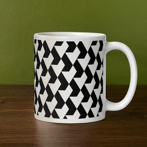 May include: White ceramic coffee mug with a black and white geometric pattern. The design features a repeating pattern of black arrow-like shapes on a white background. The mug has a curved handle and is sitting on a wooden surface.