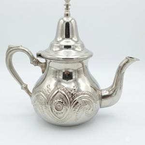 May include: A polished silver-tone metal teapot featuring an ornate embossed floral and leaf pattern around the rounded body. The teapot has a curved handle, a long slender spout, and a removable lid with a small decorative finial on top.