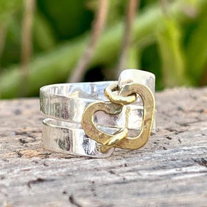 May include: A silver ring with a gold heart charm. The ring features a double band design and a hammered texture. The gold heart charm is attached to the ring with a small loop. The ring is displayed on a wooden surface.