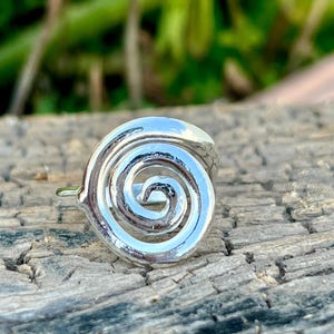 May include: A silver ring with a spiral design. The ring's spiral is centered and has a smooth, reflective surface. The ring is set against a weathered wooden surface with a blurred green background.