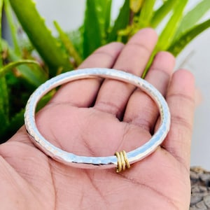 May include: A silver bangle bracelet with a hammered texture. The bracelet has a small section of gold-colored metal rings. The bracelet is shown against a background of green leaves.