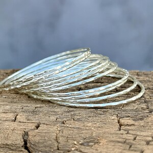 May include: A stack of silver bangles with a hammered texture. The bracelets are displayed on a weathered wooden surface, with a soft blue background. The bangles are thin and have a shiny, reflective surface.