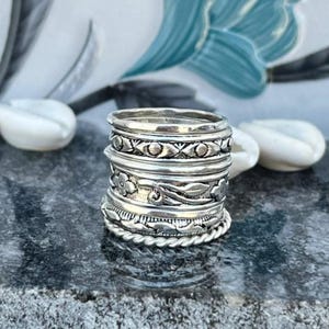 Peut inclure: Une pile de bagues en argent avec divers motifs, notamment des motifs floraux, des bordures perlées et des formes géométriques. Les bagues sont présentées sur une surface sombre et texturée, mettant en valeur leurs détails complexes et leur savoir-faire. Les bagues sont un mélange de textures et de styles.
